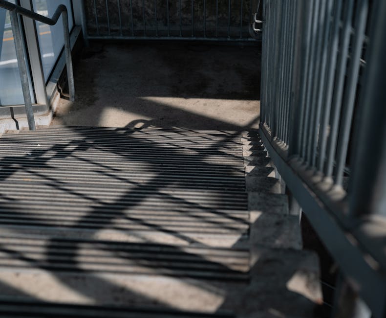 Close-up view of an outdoor metal staircase with cast iron grating steps, shaded by shadow patterns from the railing and nearby structures. The staircase is part of a residential building entrance, fe