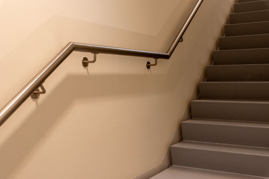 Close-up view of an outdoor metal staircase with cast iron grating steps, shaded by shadow patterns from the railing and nearby structures. The staircase is part of a residential building entrance, fe