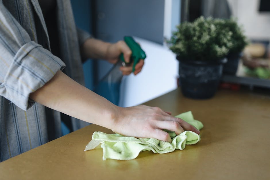 A professional cleaner in black attire with black gloves is performing surface cleaning on a wooden dining table in a modern bedroom setting. The table has a smooth, polished finish and is situated be